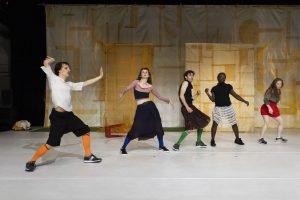 Cinq interprètes sont en train de danser, la majorité en jupe et chaussettes hautes. Derrière eux, une grande toile blanche avec des découpes géométriques jaunes."